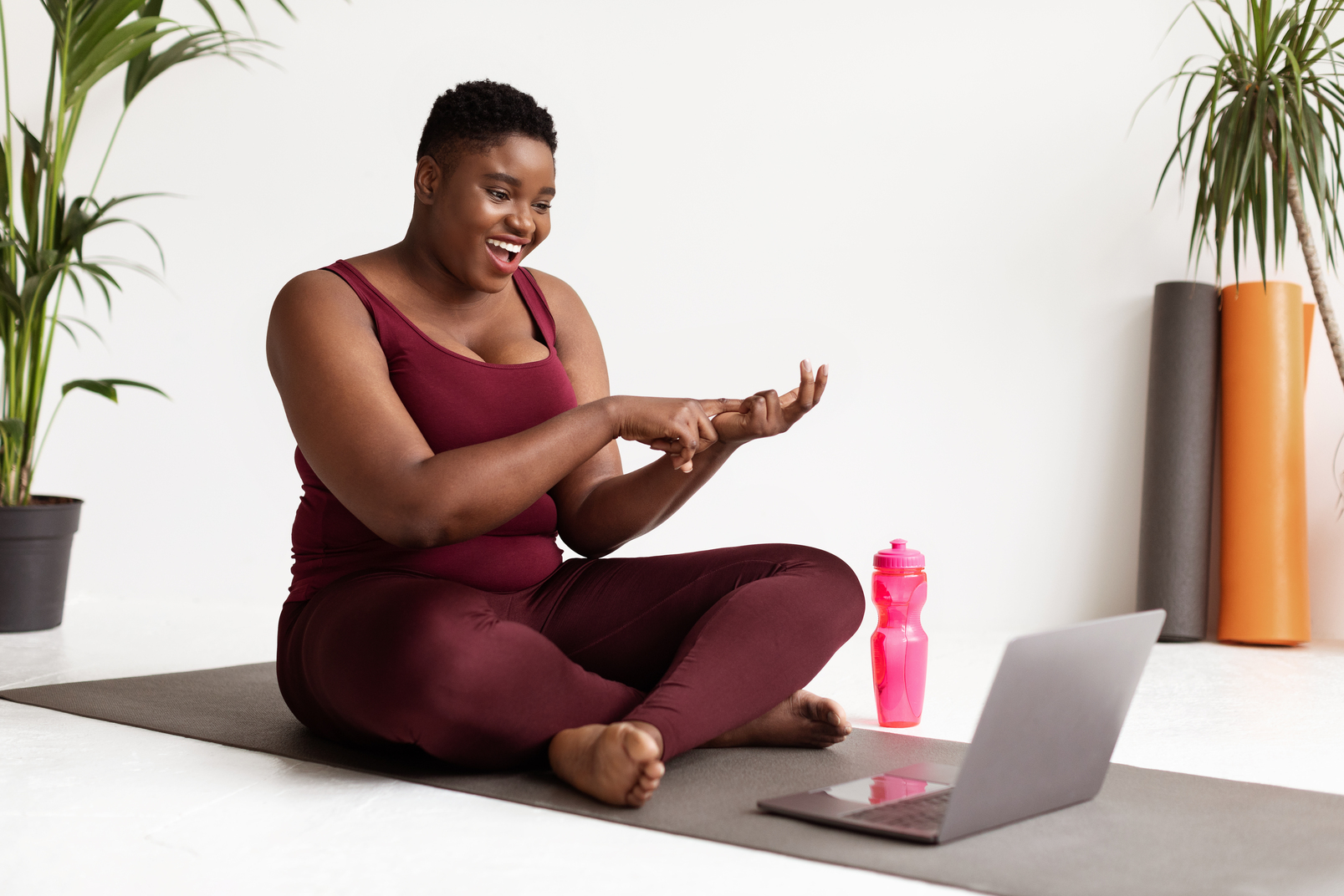 Cheerful plump millennial black woman with short hair yoga coach streaming from fitness studio, sitting on yoga mat with laptop and bottle of water, having video call with her students, copy space